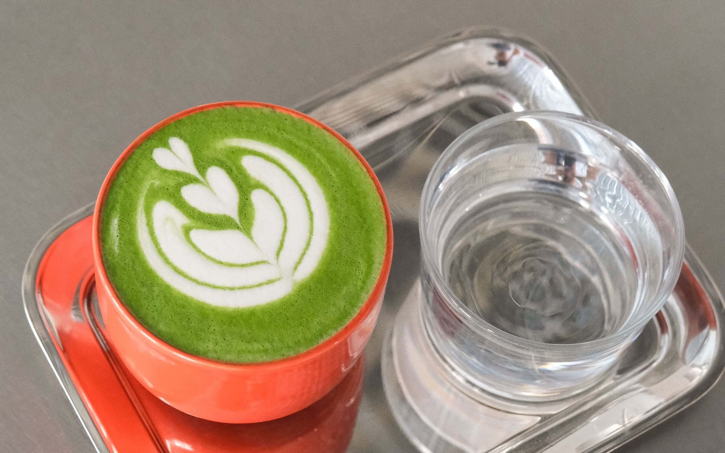 Bright green matcha latte with artistic milk foam design in an orange ceramic cup, served with a glass of water.