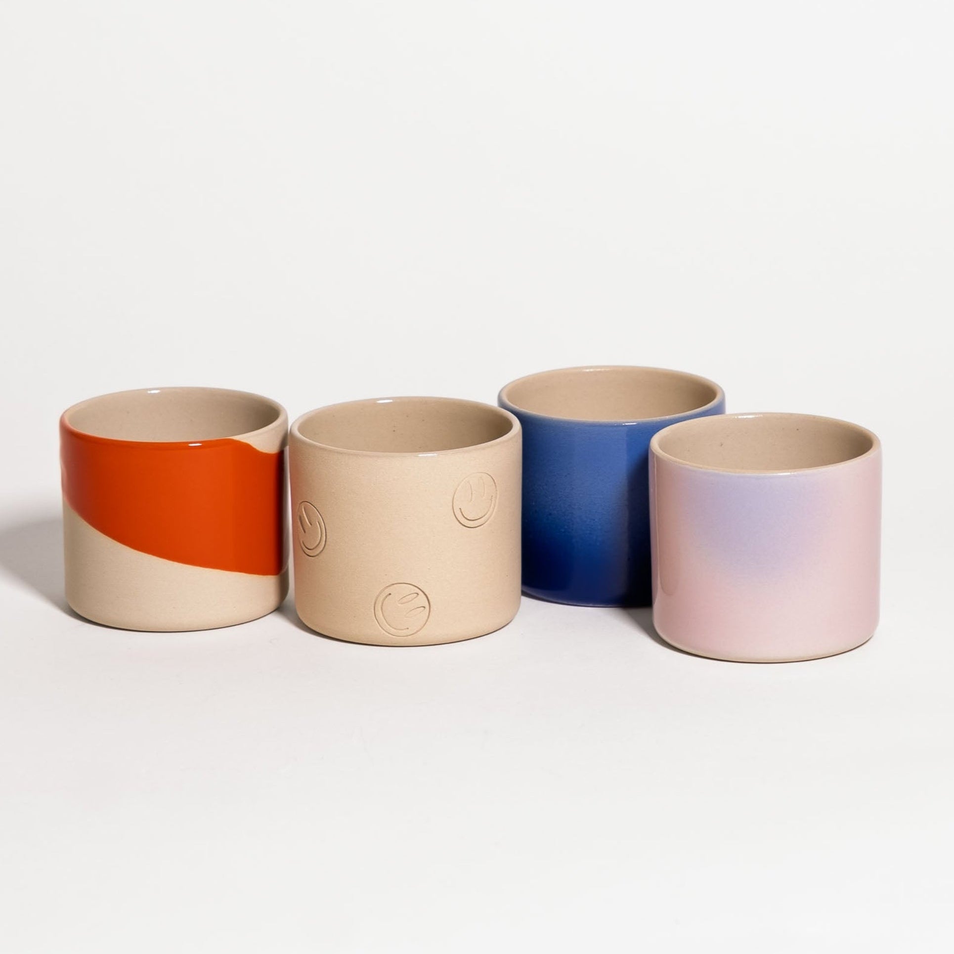 Four small cylindrical ceramic cups or candle holders in a studio setting, arranged in a row against a clean white background. The cups have a matte finish and different color designs: one is split horizontally with an orange top half and a beige bottom half; one is solid beige with embossed moon-like patterns; one is solid blue; and one has a soft pink and beige gradient.
