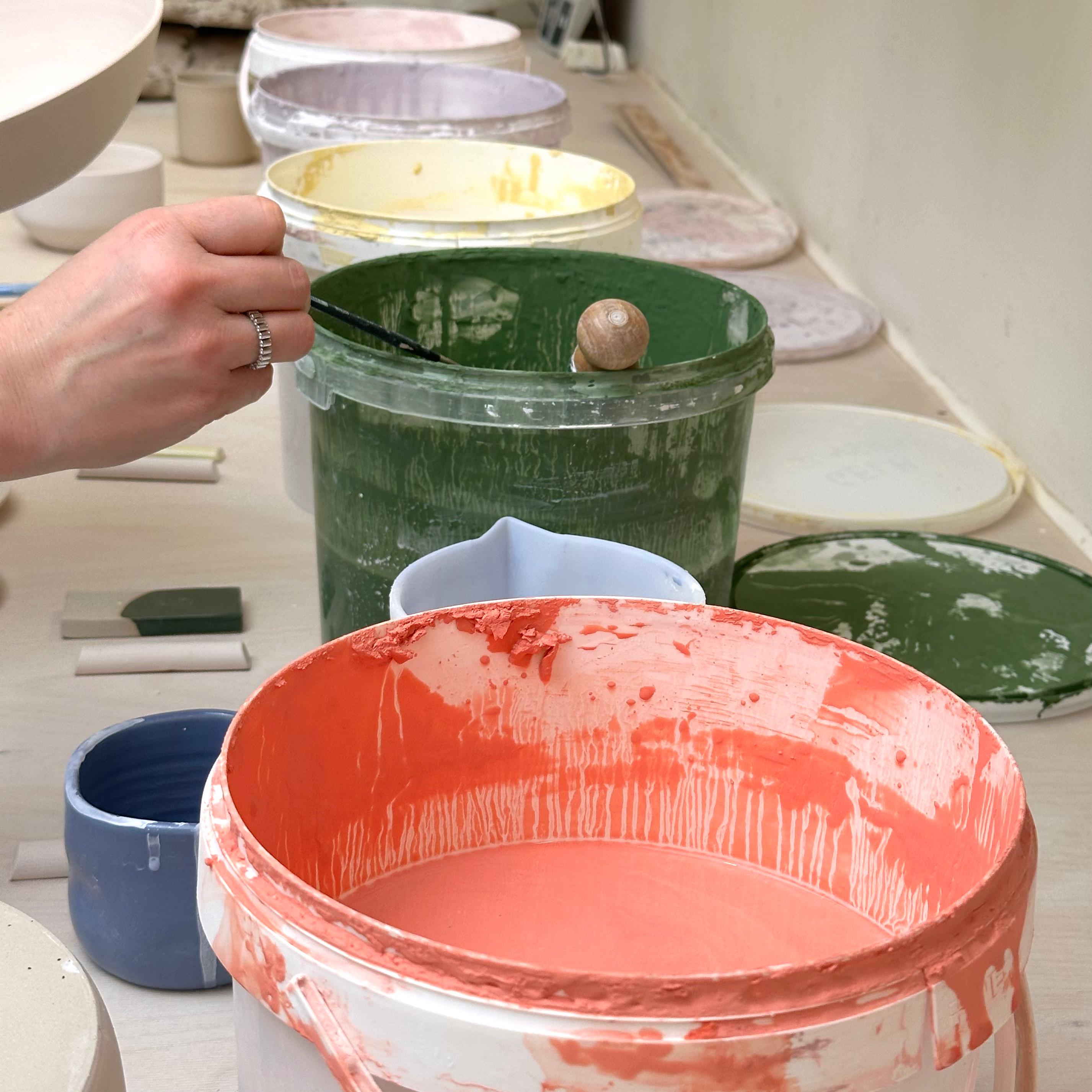 Frau nimmt mit Pinsel Glasur aus einem Eimer für den Glasur Workshop Frankfurt.