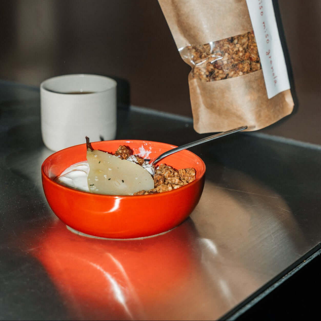 Rote handgefertigte Granola Schale mit Joghurt und Granola auf einem Tisch, daneben Tasse Kaffee.