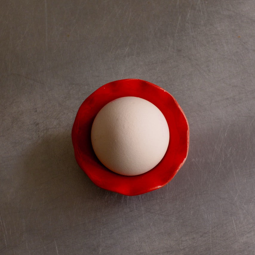 Handgemachter Eierbecher aus Steinzeug mit einem Ei in kräftiger roter Glasur auf einem Tisch.