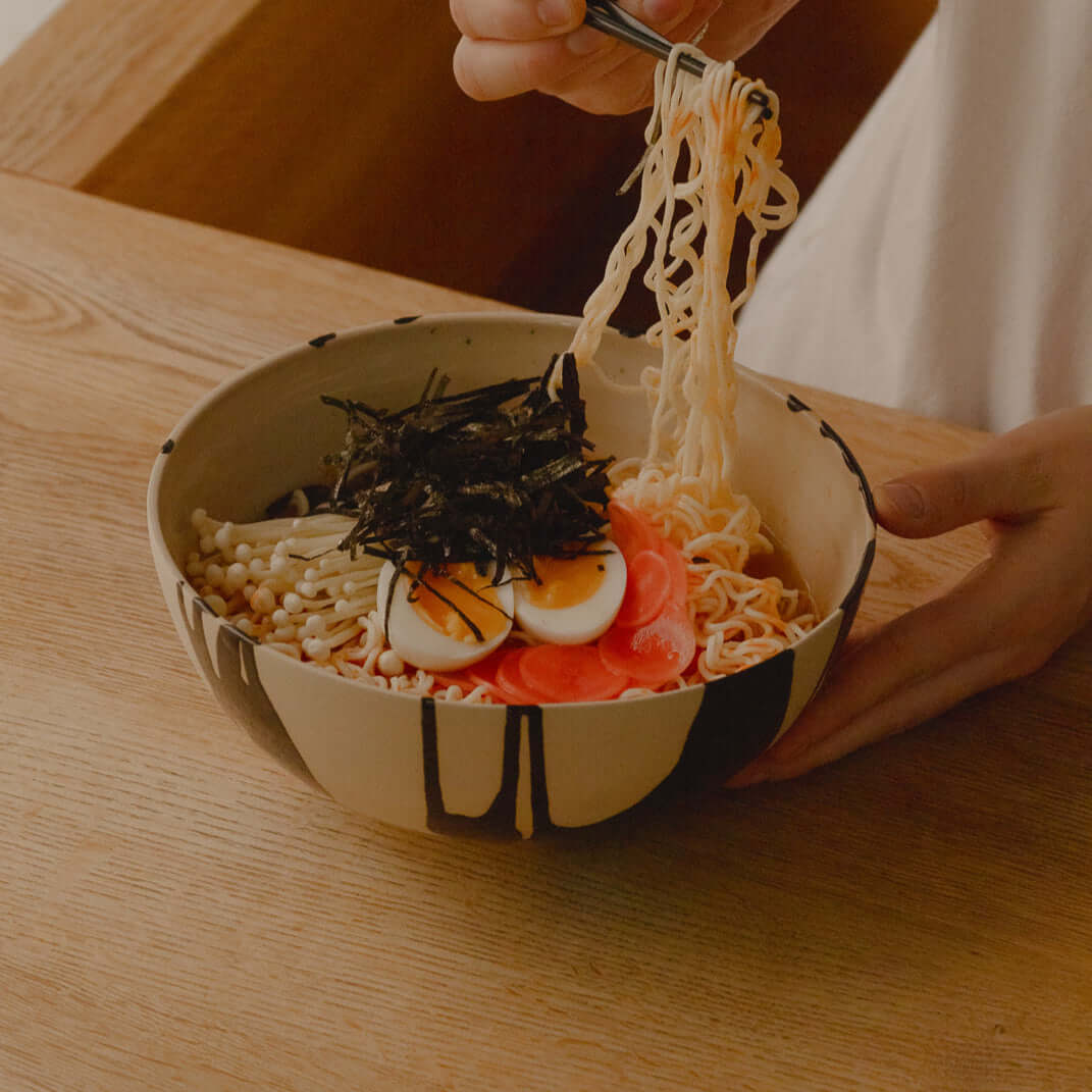 Hand holding ramen with eggs and seaweed in a stylish grey stoneware bowl beside wooden table.