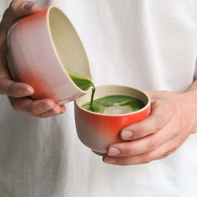 Person pouring matcha from a grey stoneware bowl with a pink-red gradient into a cup.
