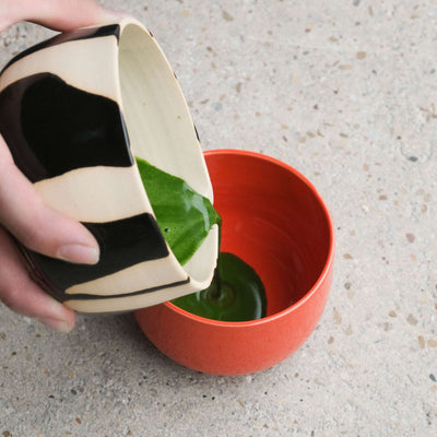 Pouring matcha from a unique serving bowl into a glossy red matcha cup made of stoneware.