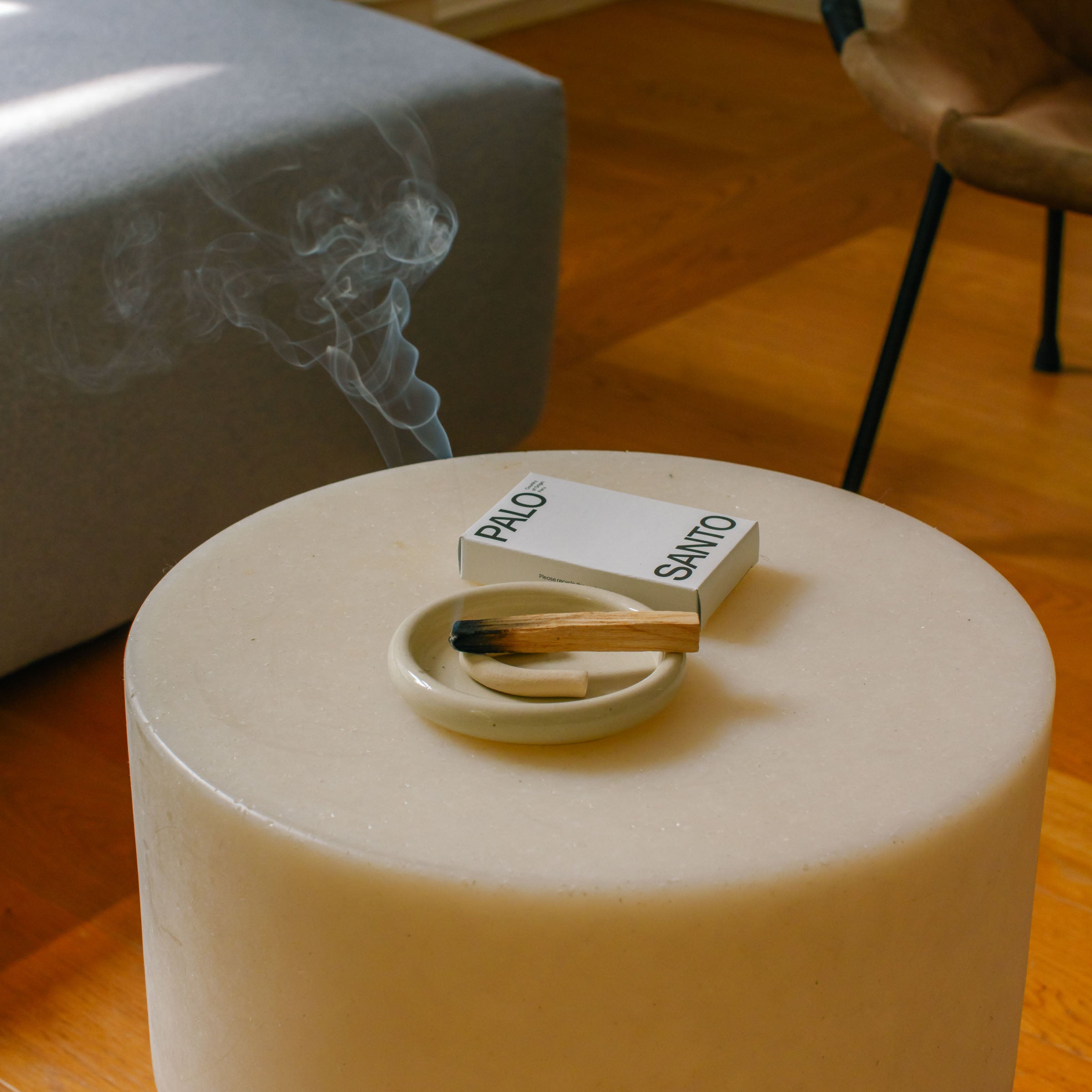 A modern living space featuring a circular stone table with a burning palo santo stick and smoke rising.