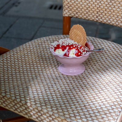 A pink dessert bowl filled with ice cream and topped with whipped cream, sprinkles, and a wafer cookie, placed on a woven rattan chair outdoors.