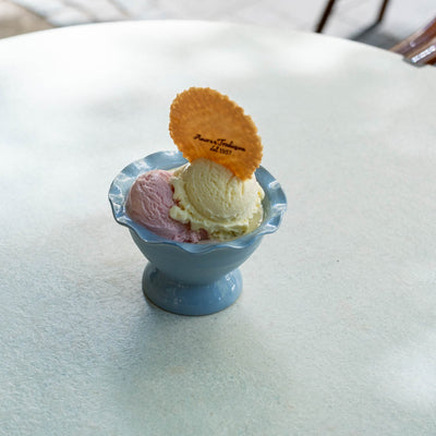 A blue ice cream sundae bowl containing pink and vanilla ice cream, topped with a round orange biscuit, set on a white outdoor table.