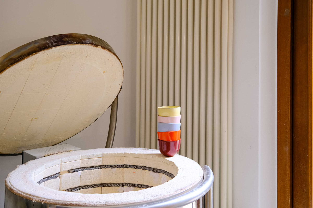A vintage ceramic stovetop kiln with its lid propped open, showing a white interior lined with dark rings, alongside a small stack of colorful glass vials on the rim, set against a background of a cream radiator and beige wall.