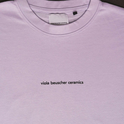 Close-up of lilac T-shirt featuring black text 'viola beuscher ceramics' on the front