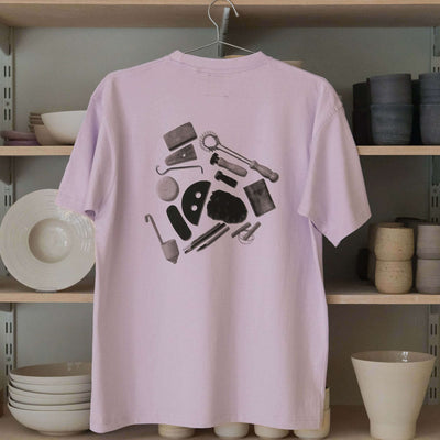 Back view of a lilac T-shirt featuring a black print of pottery tools on a shelf with pottery items.