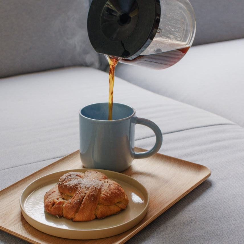 A lifestyle scene showing a coffee pour-over setup on a light wood tray. A blue ceramic mug is being filled with dark coffee from a glass pour-over coffee maker, while a golden-brown croissant sits on a beige plate in the background on a gray sofa.