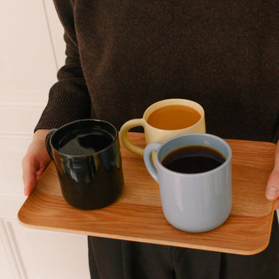 A person in a dark brown sweater holds a wooden tray carrying three ceramic mugs: one black, one pale blue-gray, and one pale yellow with coffee inside.