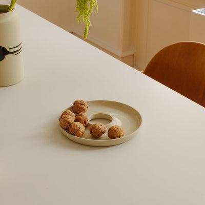 A beige ceramic donut-shaped dish holding several walnuts, placed on a white table in a softly lit interior with a chair and vase visible in the background.