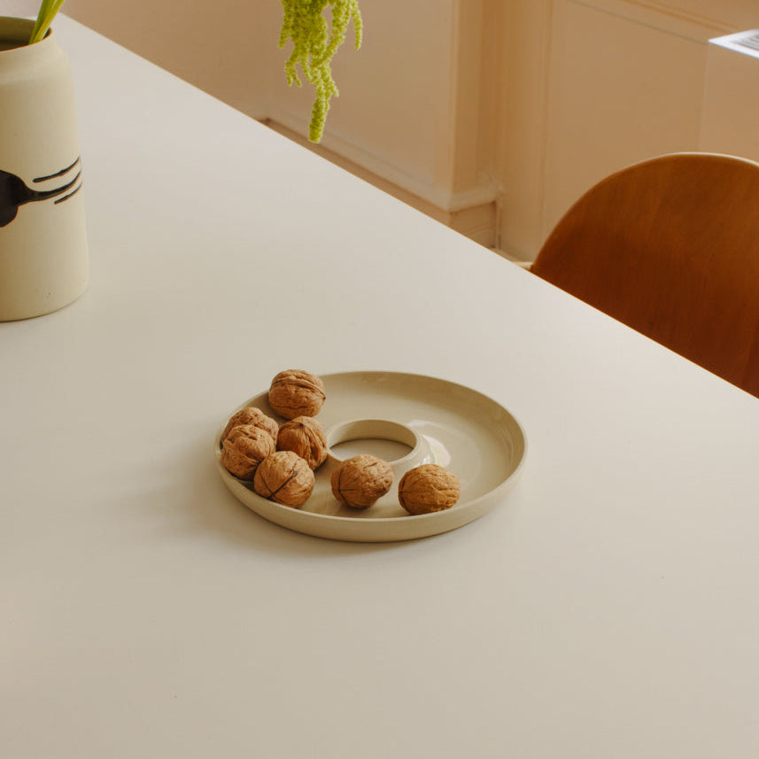 A beige ceramic donut-shaped dish holding several walnuts, placed on a white table in a softly lit interior with a chair and vase visible in the background.
