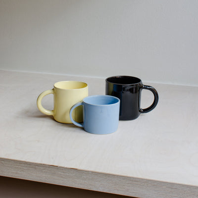 Three ceramic mugs in pastel yellow, light blue, and matte black, arranged on a light wood shelf against a neutral off-white wall.