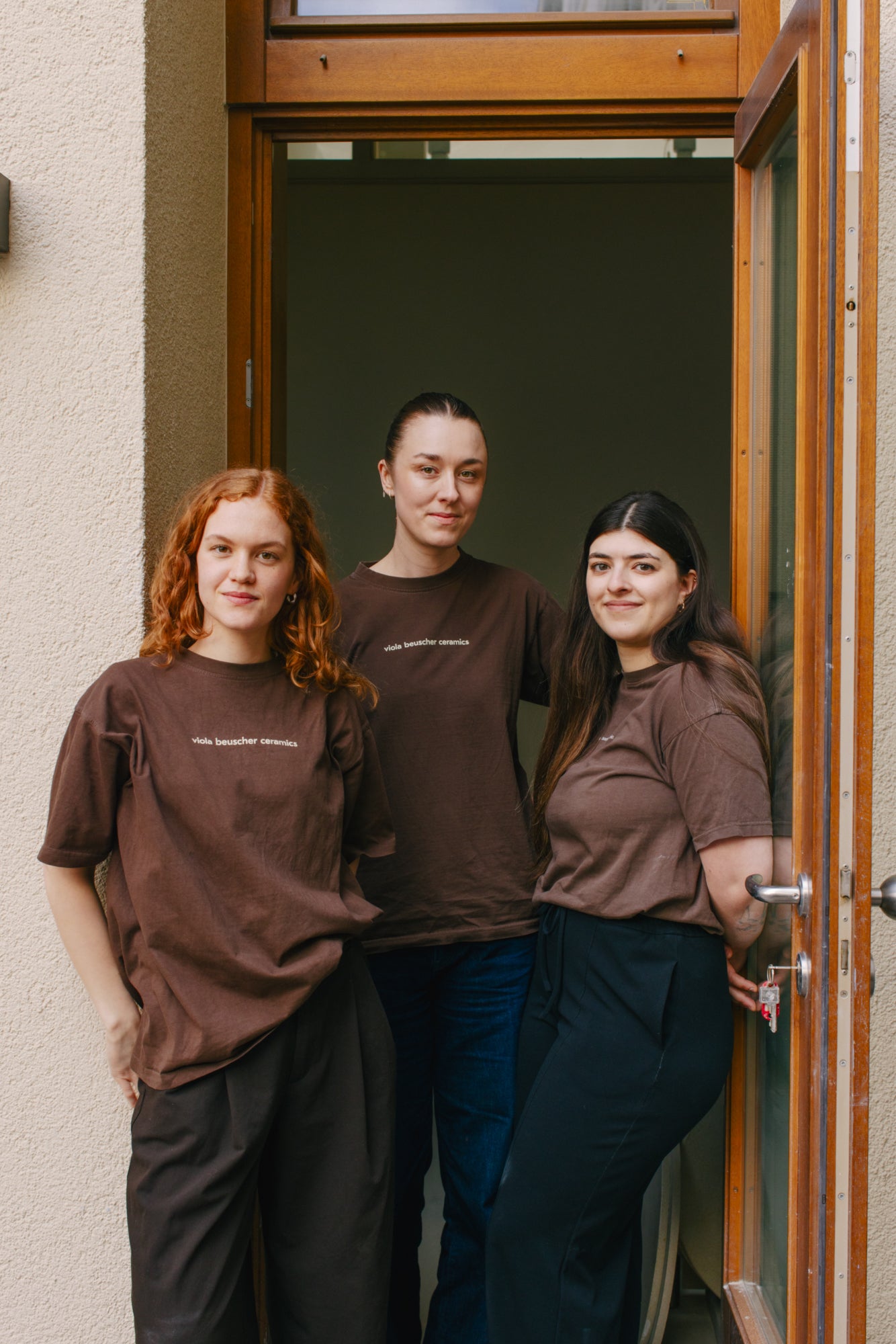 Drei Frauen in braunen T-Shirts stehen vor einer Tür, Handgemacht-Mode präsentiert.