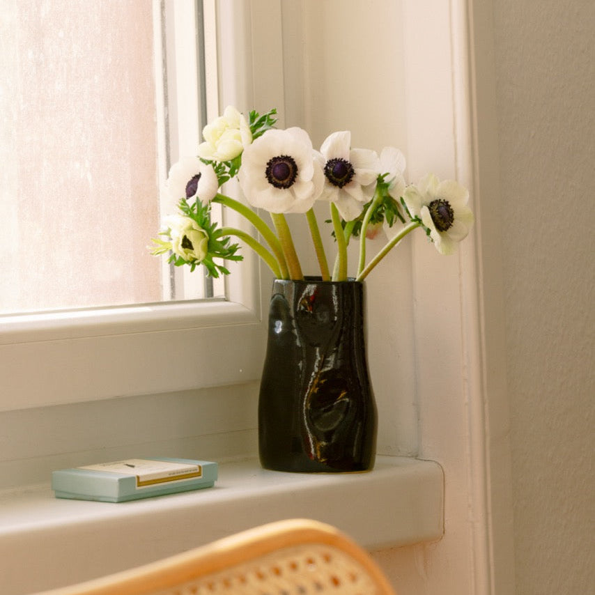 Schwarze Keramikvase mit weißen Anemonen auf einem Fensterbrett in natürlichem Licht.