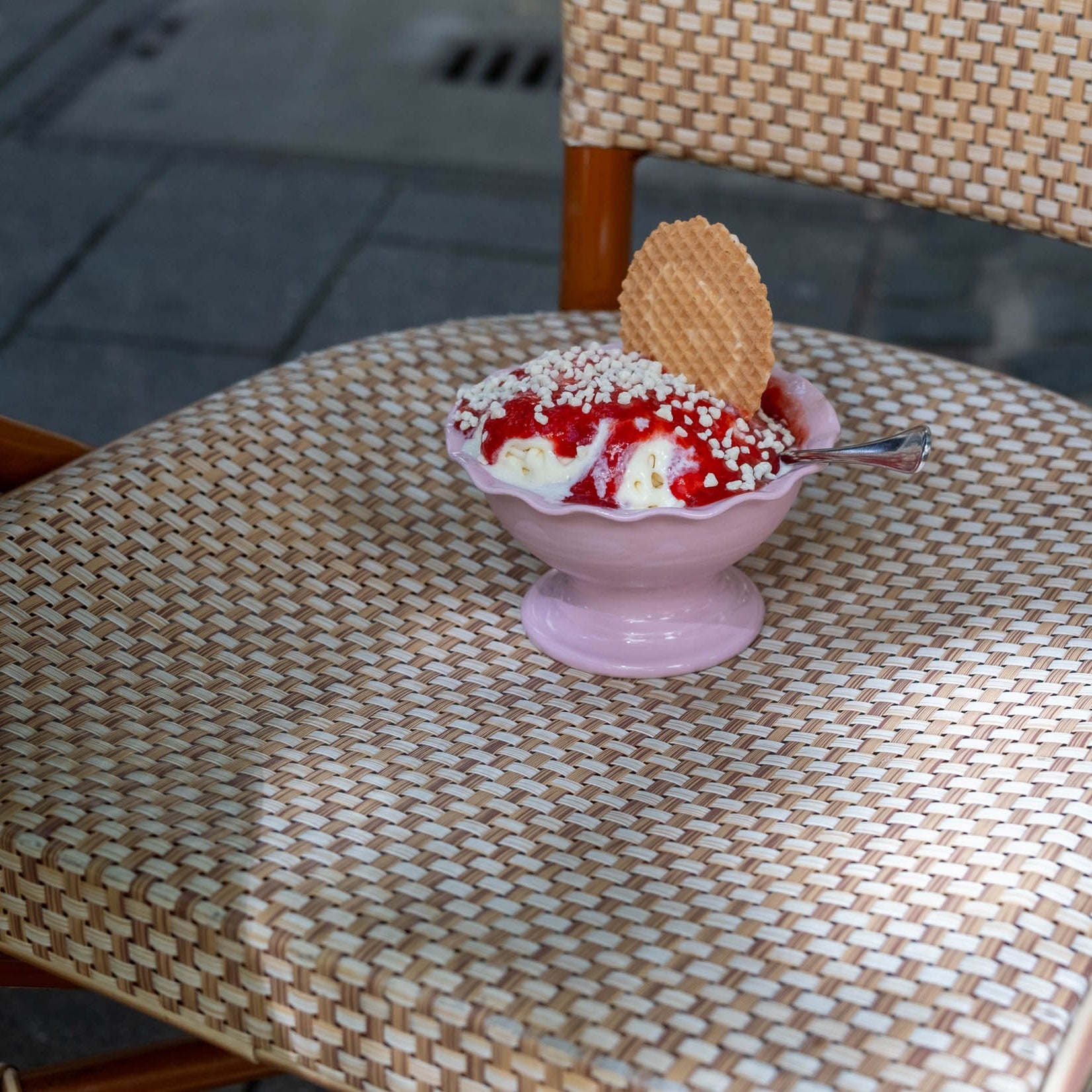 Gelato Bowl Wavy Pink mit Eis, Waffel und Schokoladensauce auf einem Stuhl aus geflochtenem Material.