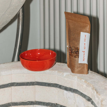 Rote handgefertigte Granola Schale neben einer Verpackung mit Müsli auf einem Steinuntergrund.
