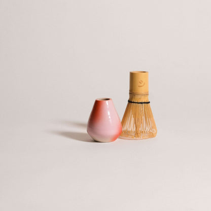 Matcha whisk holder in pink and red gradient next to a bamboo whisk, handcrafted from stoneware clay.