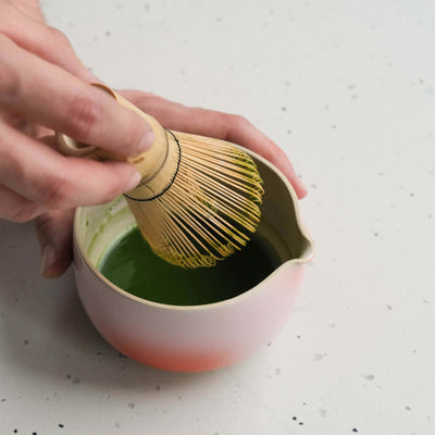 Hand holding a bamboo matcha whisk in a bowl mixing vibrant green matcha tea.