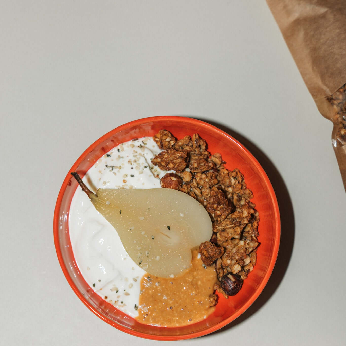 Miso Karamell Granola mit Umami serviert mit Joghurt und einer Birne in einer orangefarbenen Schüssel.