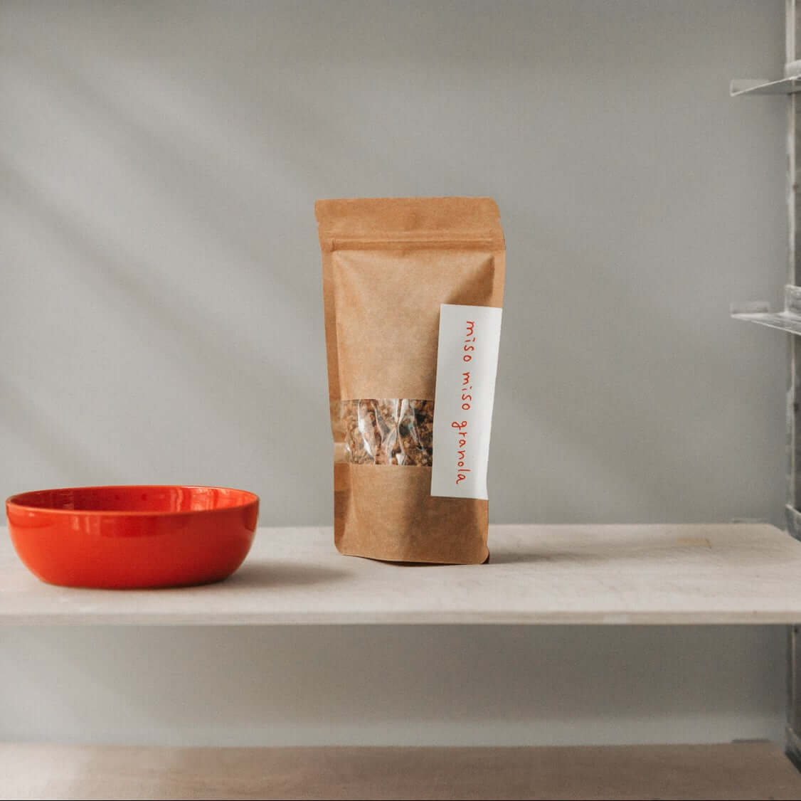 Miso-Miso-Granola - Gesalzenes Karamell & Umami in der Verpackung mit einer roten Schüssel im Hintergrund, miso karamell granola mit umami.