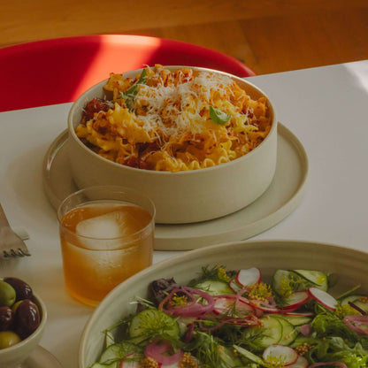 Pasta Bowl – Nomi Dark Sage filled with pasta, garnished with cheese, alongside a salad and a drink on a dining table.