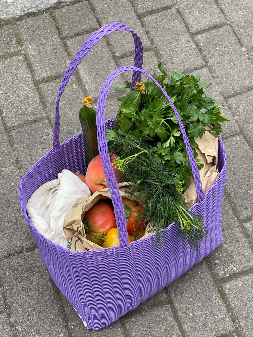 Lila Einkaufskorb mit frischem Gemüse und Kräutern auf einem Gehweg.