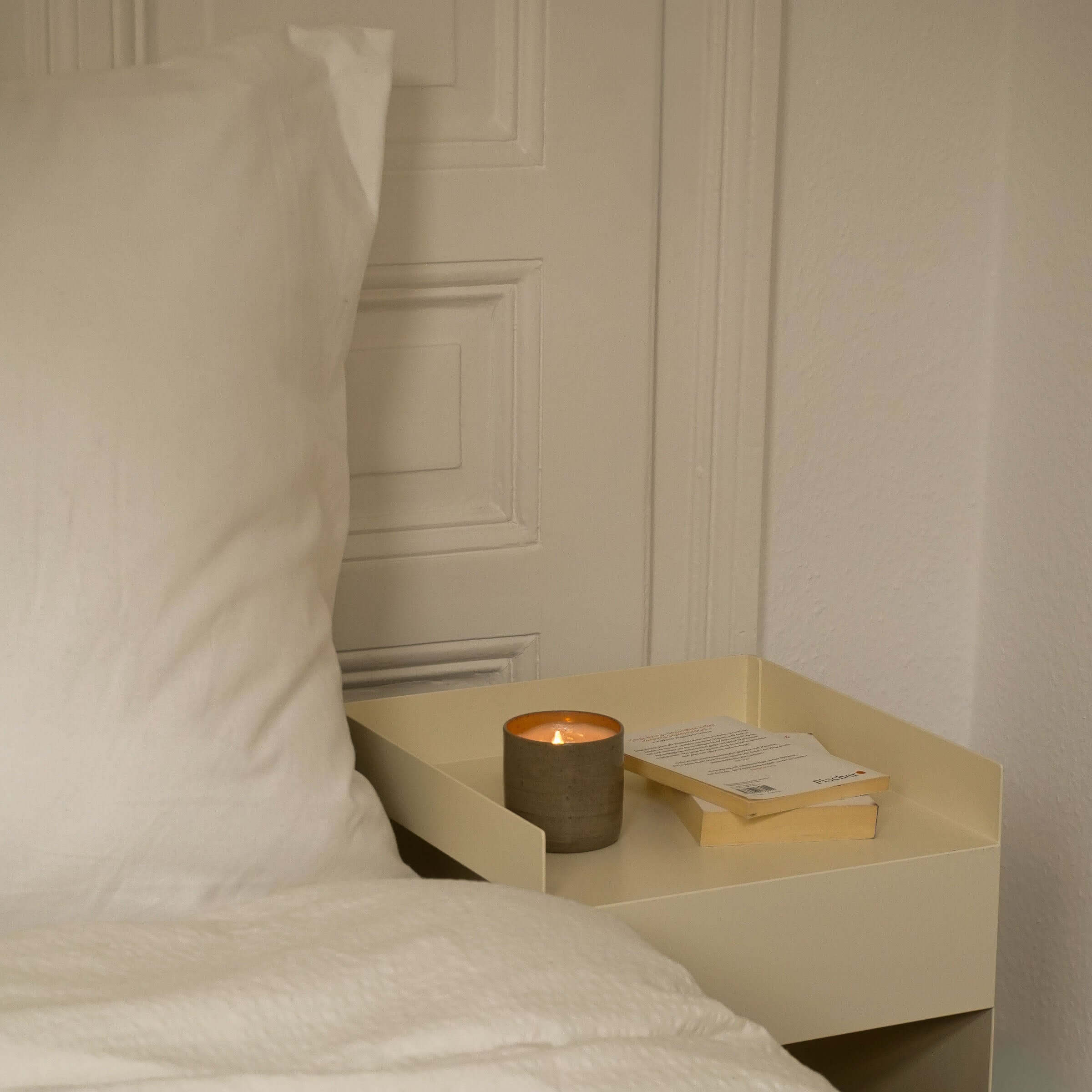Duftkerze Sandelholz auf einem Nachttisch neben einem Kopfkissen und einem Buch, sanfte Beleuchtung.