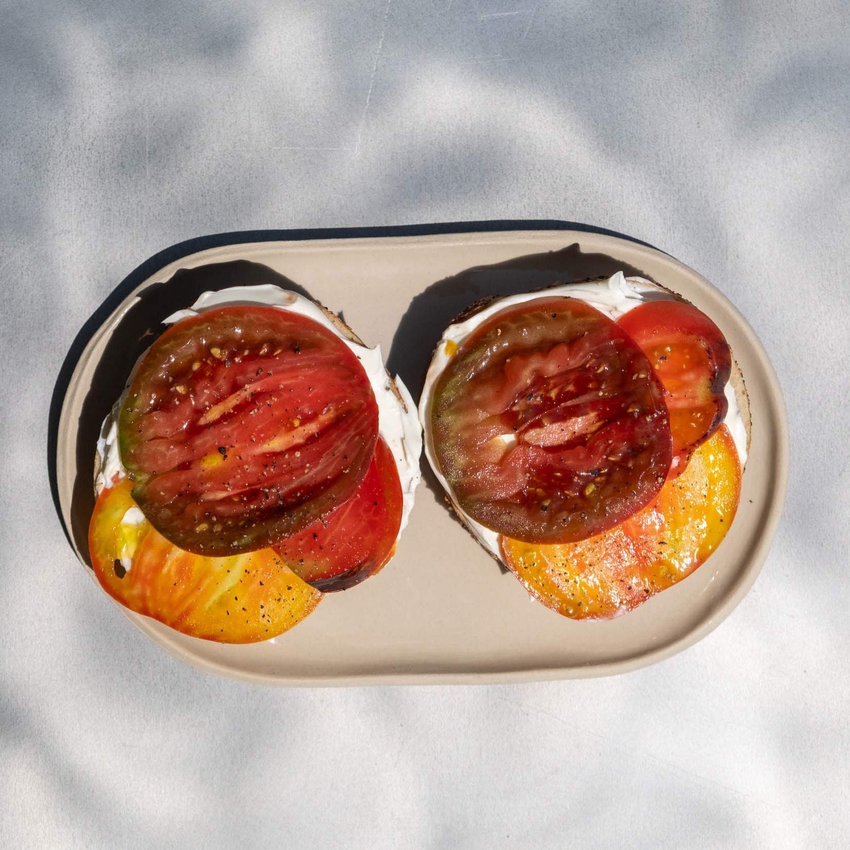 Two halves of an oval white plate with a black rim, filled with sliced red and yellow tomatoes, set against a white background with dappled shadows.