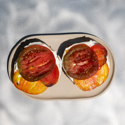 Two halves of an oval white plate with a black rim, filled with sliced red and yellow tomatoes, set against a white background with dappled shadows.